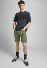 Camiseta a rayas en azul marino y negro, pantalones cortos verdes arremangados, calcetines oscuros y zapatillas grises con detalles blancos. Ajuste casual, tejido ligero.