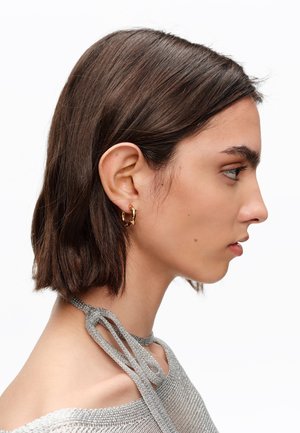 Mujer joven con cabello corto y oscuro, luciendo pendientes de aro dorados y un top plateado de punto con tirantes atados, mostrada de perfil sobre un fondo blanco.