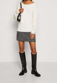 Witte geribbelde trui, zwart-wit geblokte rok en zwarte leren laarzen tot de knie. Model draagt een kleine zwarte handtas.