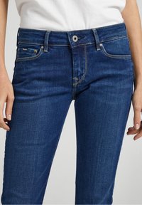 Mörkblå denimjeans med en slät yta, femfickorsdesign och silverdetaljer med synliga sömnadsdetaljer vid sömmarna.