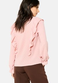 Pink blouse with ruffled accents, dotted texture, long sleeves with elastic cuffs, paired with brown pants. Simple, lightweight design.