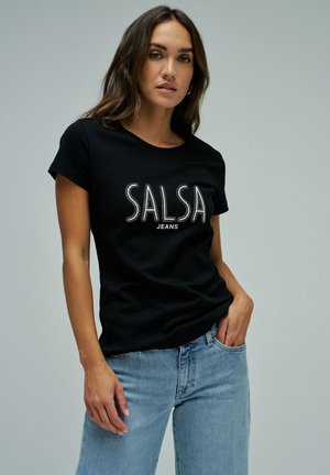 Mujer joven con camiseta negra "SALSA JEANS" y vaqueros azul claro, de pie con una mano en el bolsillo sobre un fondo liso.