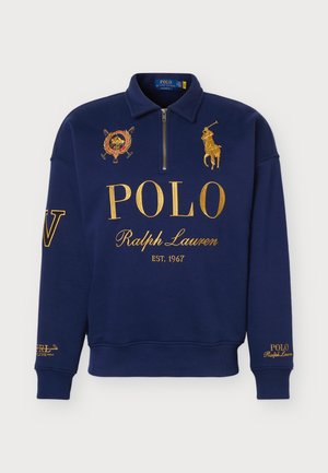 Un homme porte un sweat-shirt bleu marine Polo Ralph Lauren à fermeture éclair courte, avec des lettres dorées, un jean délavé clair et des baskets blanches sur un fond uni.
