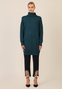 Teal strikket turtleneck tunika, oversized pasform, lange ærmer, parret med sorte jeans med frynser og perle detaljer ved kanten, sorte hæle.