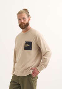 Sudadera beige de manga larga, cuello redondo y un bolsillo de parche azul marino con texto amarillo. Combinada con pantalones verdes. Textura suave.