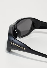 Black sunglasses with a shiny finish, featuring a bold gold "Oakley" logo on the frame. Dark lenses with a streamlined, sporty design.