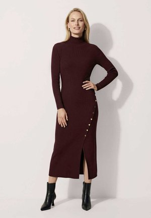 Femme debout portant une robe midi bordeaux foncé à manches longues avec des boutons sur le côté et une fente à l'avant, associée à des bottines noires.