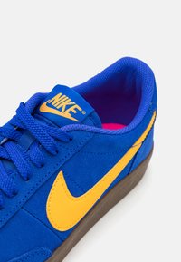 Sapatilha azul de camurça com um logo da Nike amarelo, língua de malha texturizada e sola de borracha castanha. Apresenta atacadores azuis e interior acolchoado.