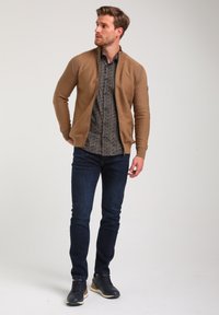 Cárdigan de punto marrón con cuello alto, llevado sobre una camisa de estampado, combinado con jeans oscuros y zapatillas negras. Diseño de atuendo casual.