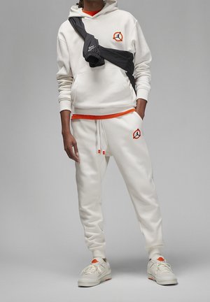 Osoba ubrana w biały hoodie i spodnie dresowe z pomarańczowymi akcentami i logo Jordan, czarna torba na ramię oraz białe trampki.