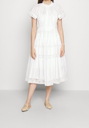 Mujer con un vestido midi blanco con encaje, mangas cortas y zapatos beige sin cordones, de pie frente a un fondo claro y liso.