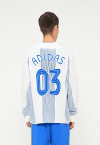 Bela pletena majica Adidas z modrim številom "03" in napisom "ADIDAS". Oblikovanje vključuje pikčasto teksturo in vertikalne črte.