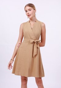 Beige wrap dress with a v-neckline, sleeveless design, and a tie belt. Made from textured material, features a flared skirt.
