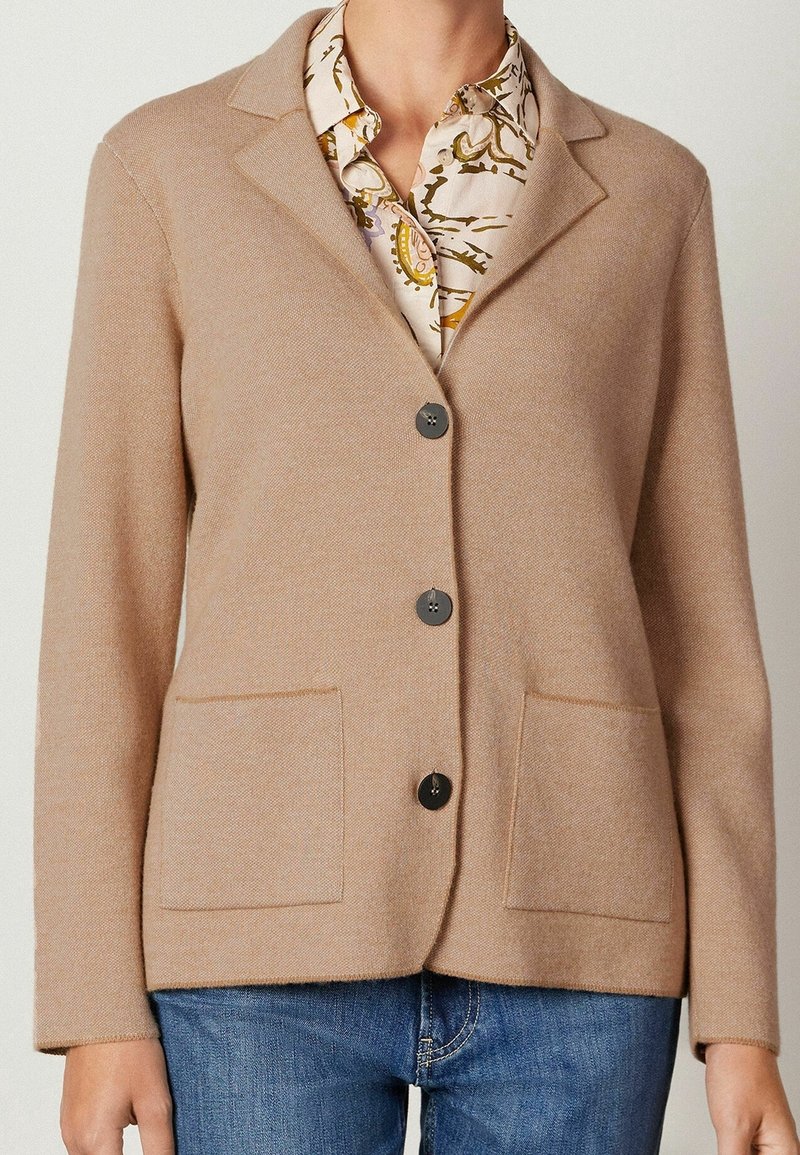 Blazer beige boutonné avec deux poches avant porté sur une chemise à motifs et un jean bleu, montré du cou aux hanches.