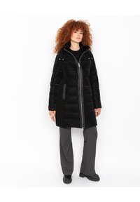 Abrigo acolchado negro con cierre de cremallera, cuello alto y bolsillos laterales. Presenta un material brillante, un ajuste relajado y detalles contrastantes en gris.