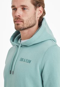 Mint green hoodie with drawstrings, featuring "EBB & FLOW" logo in darker green and orange. Made of soft, textured fabric.
