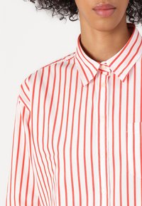 Button-up shirt with vertical red and white stripes, collar, and pocket on the left. Made from smooth fabric with subtle sheen.