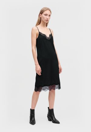 Femme portant une robe noire sans manches avec un bord en dentelle et des bottines noires, debout devant un fond gris clair uni.