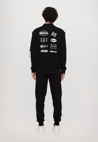 Black sweatshirt and pants set. Sweatshirt features various white logos on the back. Plain design and fitted cuffs. White sneakers shown.