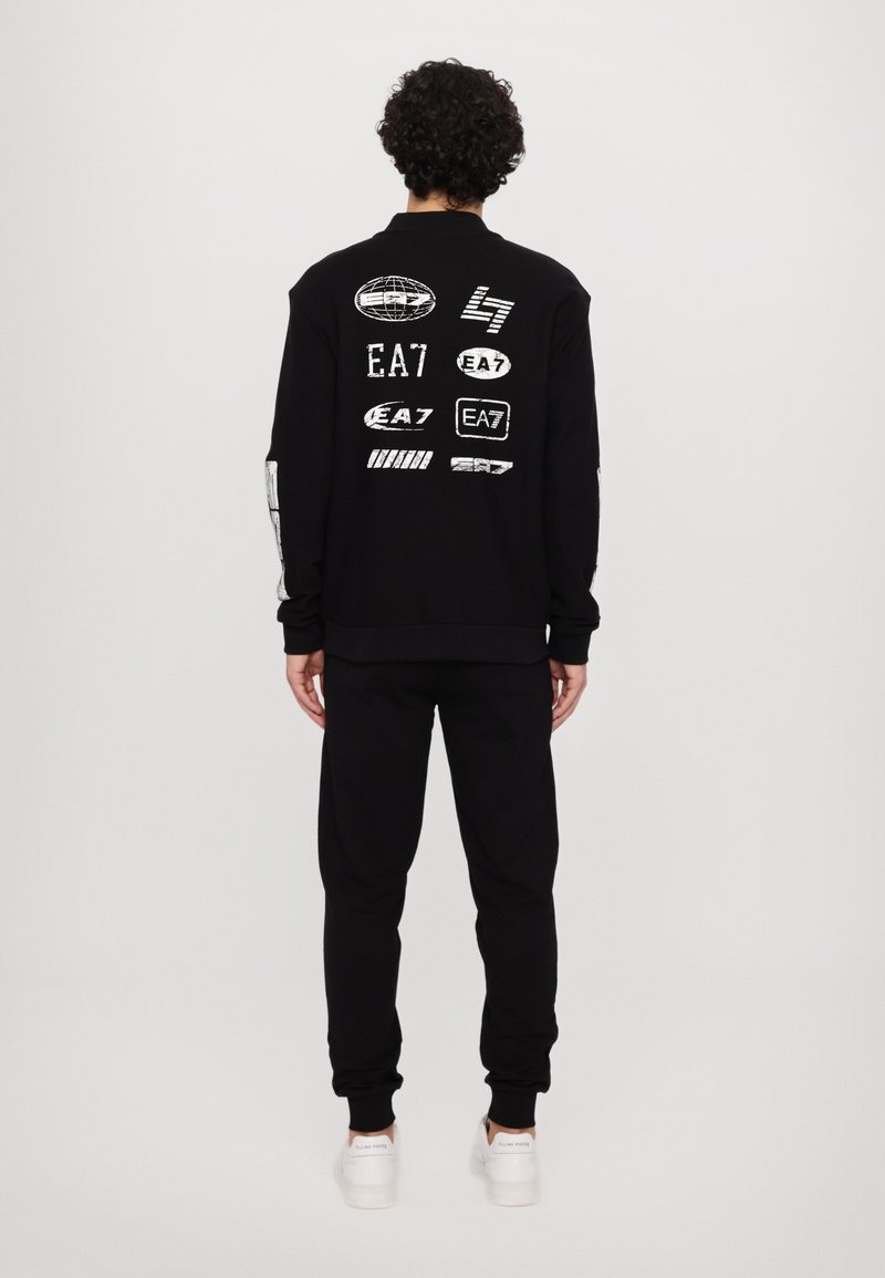 Conjunto de sudadera y pantalones negros. La sudadera presenta varios logotipos blancos en la parte posterior. Diseño sencillo y puños ajustados. Se muestran zapatillas blancas.