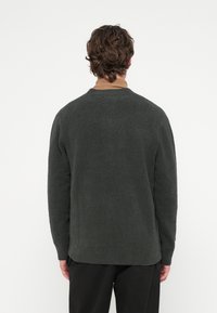 Sötétzöld pulóver puha anyaggal, lekerekített nyakkivágással és hosszú ujjal. Bordázott szegély és mandzsetta, barna galléros ing fölött viselve.