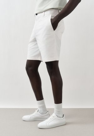 Homme portant un short blanc, des chaussettes blanches et des baskets blanches, debout avec une main dans la poche sur un fond uni.