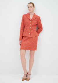 Blazer en tweed rouge avec de grands boutons et un design à péplum, associé à une jupe adaptée assortie. Le matériau a une finition texturée.