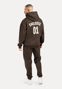 Felpa marrone con stampa "SMILODOX 01" sul retro, abbinata a pantaloni tuta coordinati e sneakers bianche. Texture morbida, vestibilità rilassata.