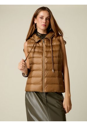 Giovane donna con capelli lunghi indossa un gilet imbottito marrone senza maniche sopra un top a quadri e una gonna lucida verde oliva, tenendo il cordino del gilet.