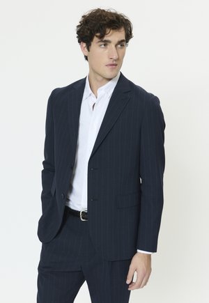 Marineblå jakkesæt med nålestriber, to frontlommer, en hakke krave og to knapper. Båret over en hvid skjorte. Tekstureret stof.
