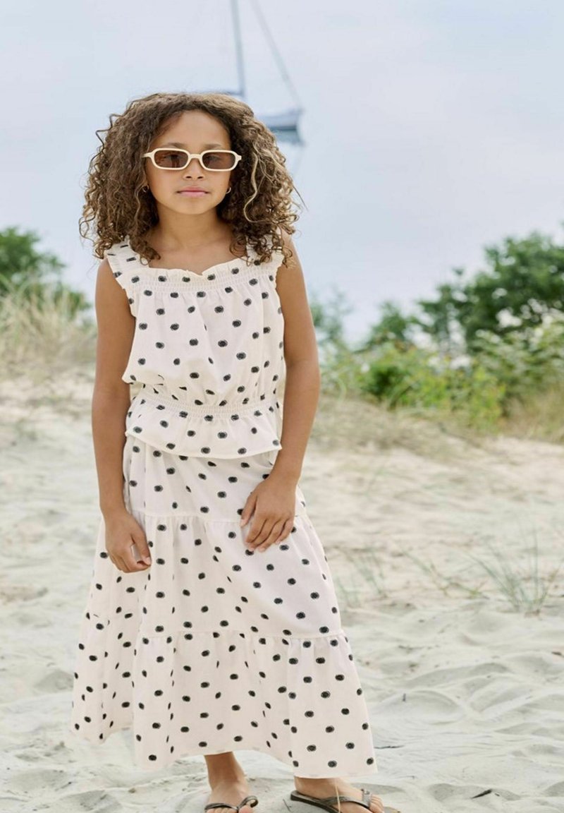 Fille aux cheveux bouclés portant des lunettes de soleil blanches et une robe blanche à pois noirs, debout sur une plage de sable avec de la verdure en arrière-plan.
