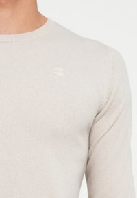 Sweater de malha cinza claro com gola redonda, apresentando um logo bordado discreto no peito. Textura suave e design ajustado.