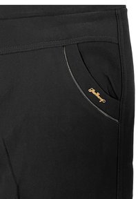 Schwarze Hosen aus weichem Stoff verfügen über eine geschwungene Tasche mit einem goldenen Logo. Der glatte Stoff hat dekorative Nähte entlang des Taschensaums.