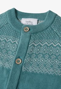 Cardigan en tricot teal avec un col rond, décoré d'un motif géométrique blanc et de boutons beige. L'étiquette indique "Stellow & Friends."