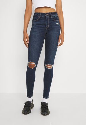 Kobieta w ciemnoniebieskich, podartych obcisłych dżinsach, białym krótkim topie, białych skarpetkach i czarnych mokasynach na jednolitym jasnym tle.