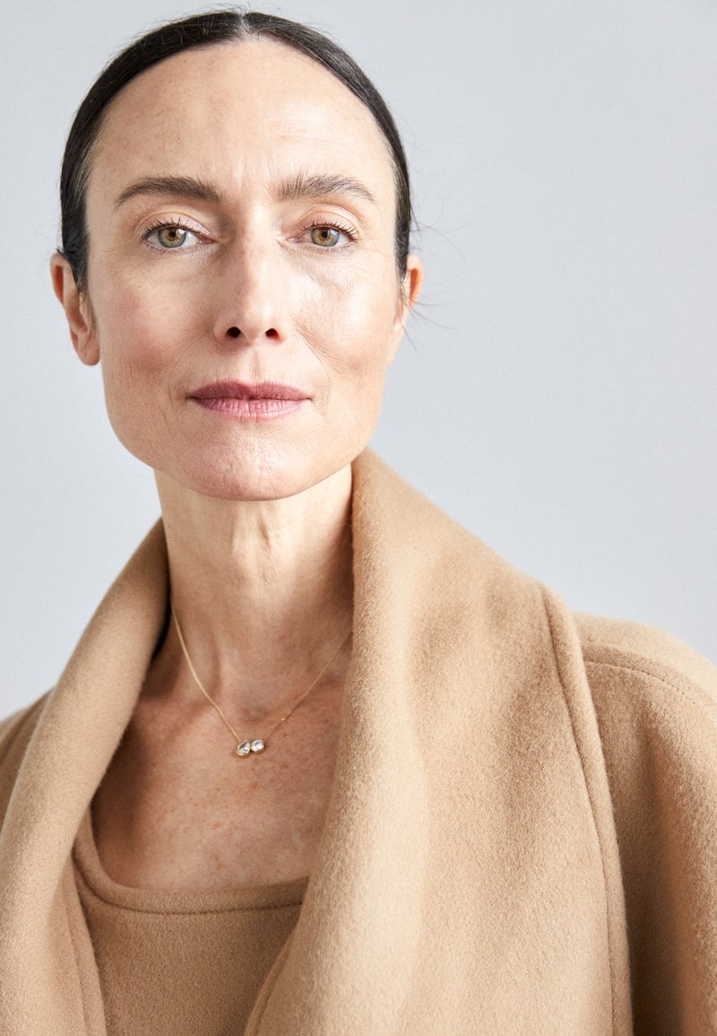 Bežinis vilnonis paltas su plačia apykakle, uždėtas ant atitinkančios palaidinės; švelnus auksinis kaklo papuošalas, papuoštas dviem mažais akmenimis. Neutralus fonas.