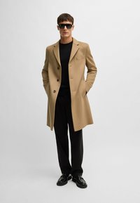 Cappotto di lana color tan con revers, tre bottoni e taschino. Indossato sopra un maglione nero e abbinato a pantaloni e scarpe nere.