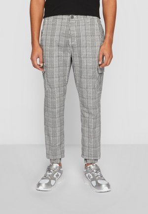 Homme portant un pantalon cargo à carreaux gris et blancs et des baskets argentées, debout devant un fond uni.