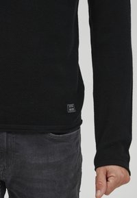 Zwarte gestructureerde trui met lange mouwen, geribbeld biesje en een klein label nabij de zoom. Gepaard met donkergrijze jeans.