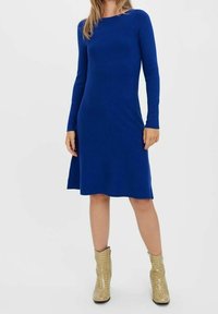 Robe à manches longues, longueur genou, de couleur bleu vif uni, en tissu doux. Associée à des bottes beiges texturées.