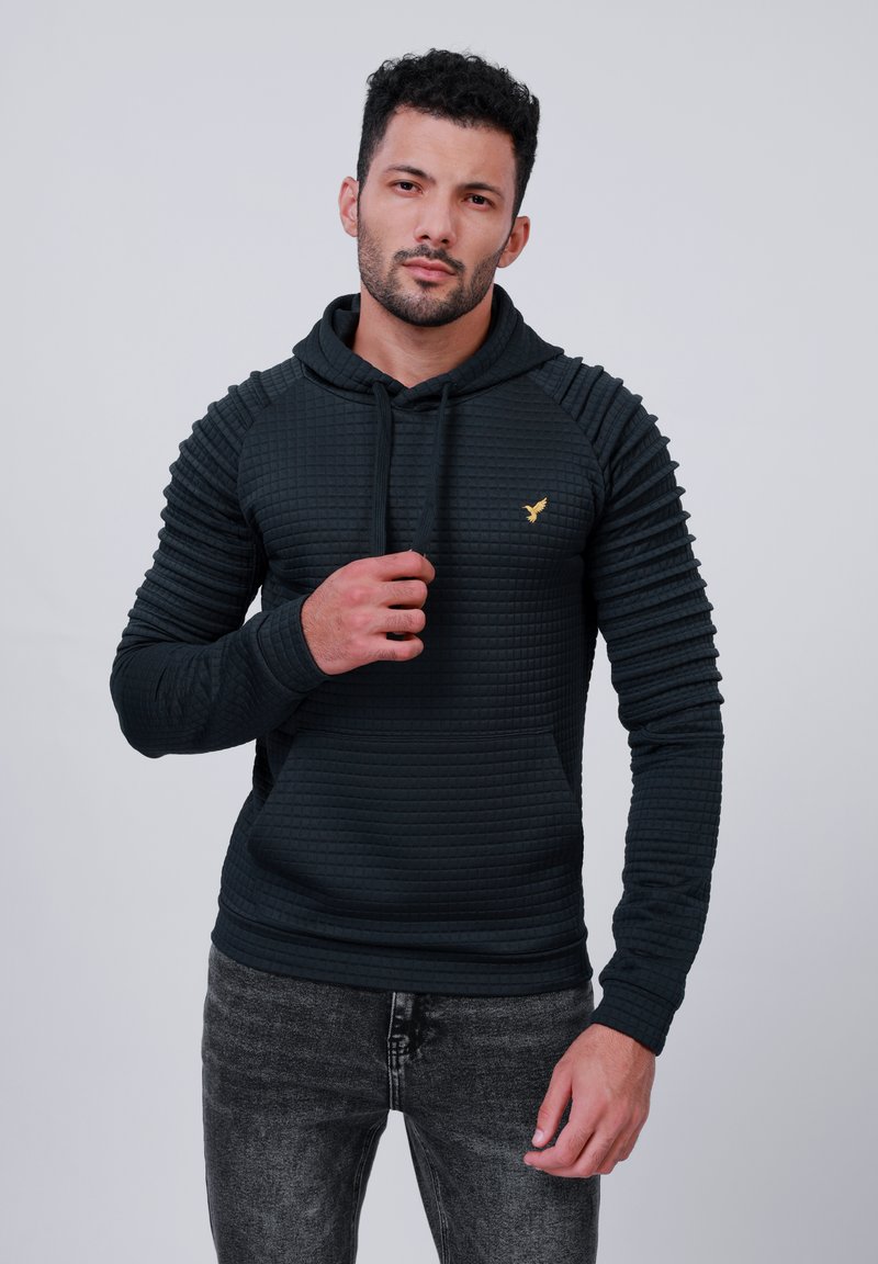 Sweatshirt negru cu glugă, cu textură matlasată, accente ribbate pe umeri și un mic logo auriu pe piept. Asortat cu blugi închiși la culoare.