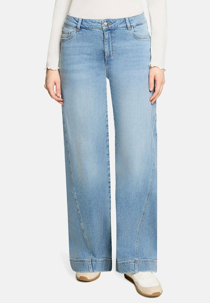 Jeans larges bleu clair portés avec des baskets blanches et un haut blanc à manches longues, montrés de la taille jusqu'aux pieds.