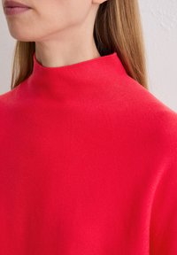 Roter hochgeschlossener Pullover aus strukturiertem Strickstoff, der eng anliegt und ein minimalistisches Design aufweist. Der Hintergrund ist neutral und unstrukturiert.