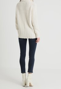 Nainen, joka on pukeutunut tekstuurilla varustettuun vaalean kermanväriseen neulepaitaan, tummiin kapeisiin farkkuihin, joissa on purjeet, ja valkoisiin korkokenkiin, seisoo selkä kameraan päin.