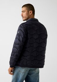 Marineblaue gesteppte Steppdaunenjacke mit abgerundetem Muster, versehen mit einem Markenabzeichen auf der Schulter und gerippten Bündchen. Legere Passform und leichtes Design.