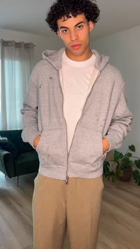 Ung mand med krøllet hår iført en grå lynlåshoodie over en hvid t-shirt og beige bukser står indendørs med hænderne i lommerne.