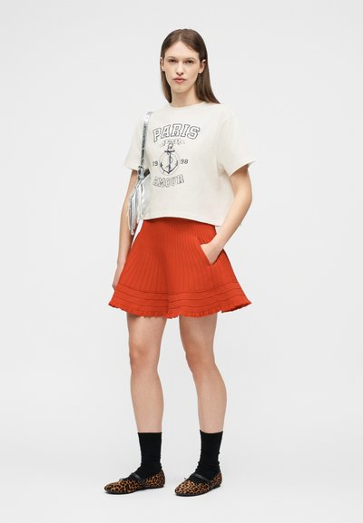 Νεαρή γυναίκα που φοράει λευκό T-shirt με στάμπα, κόκκινη πλισέ φούστα, μαύρες κάλτσες, παπούτσια με λεοπάρ μοτίβο και ασημένια τσάντα ώμου σε λευκό φόντο.