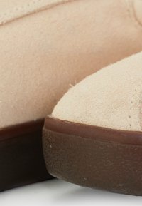 Zapato de ante beige con punta redondeada y suela de goma marrón texturizada. Detalles de costura visibles a lo largo del borde. Textura suave, diseño minimalista.