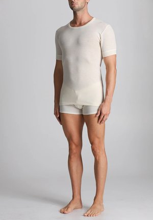 Uomo che indossa una camicia a maniche corte beige attillata e slip coordinati, poggiato a piedi nudi su uno sfondo grigio chiaro.