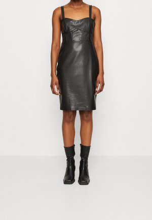 Schwarzes ärmelloses Kleid aus Kunstleder mit tailliertem Schnitt, knielangem Saum und schwarzen Knöchelboots mit quadratischer Spitze und seitlichen Reißverschlüssen.